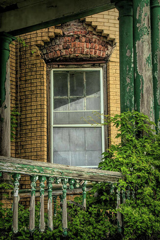 This images shows the window of the abandoned Bachelor Officers' Quarters at Fort Hancock on Sandy Hook, NJ.  The brick facade has fallen away from the top of the window revealing red brick underneath.  The railing is falling apart and has green peeling paint. Virginia creeper is growing up the porch column.